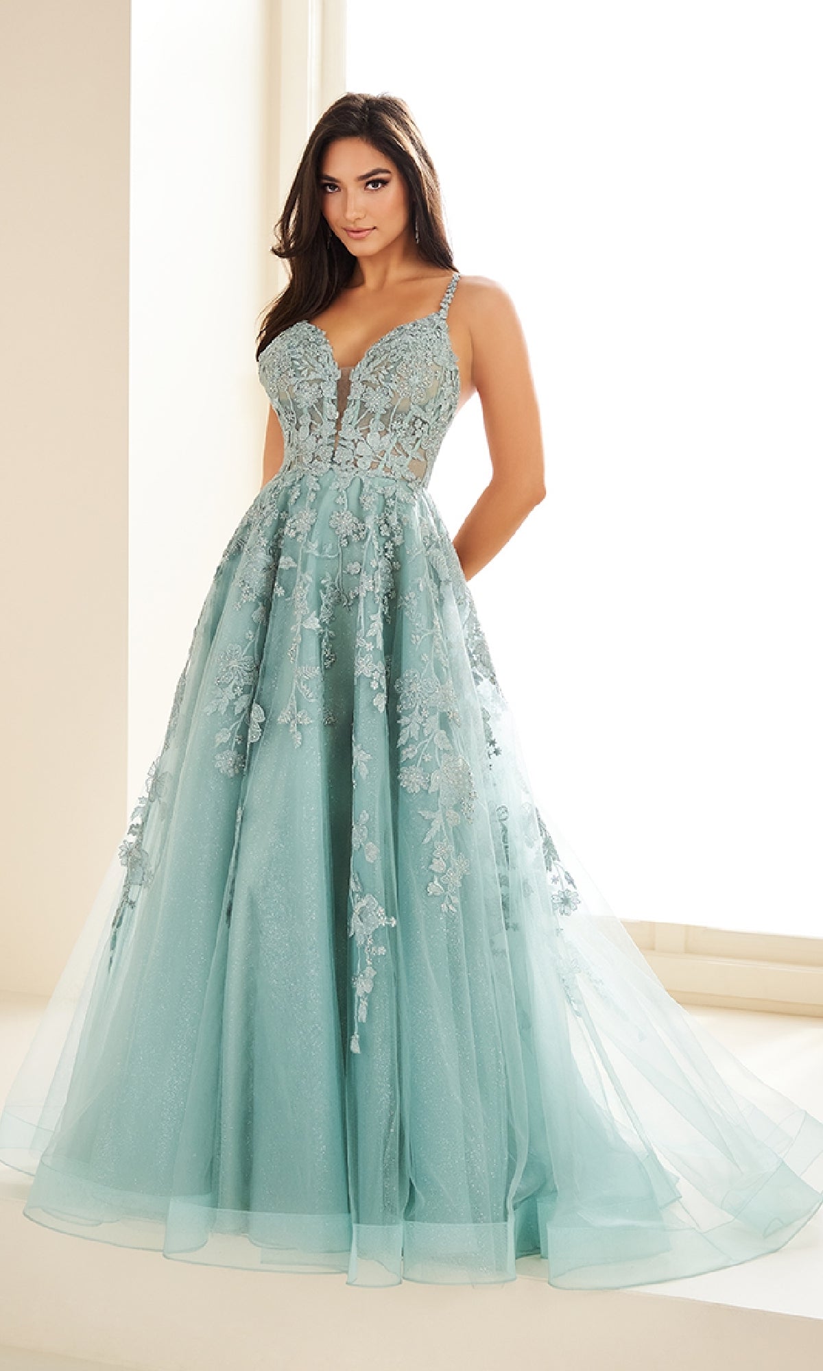 A woman models the Ellie Wilde EW36015, a light blue, floor-length prom dress featuring lace embroidery and a fitted corset bodice, posing indoors against a bright background.